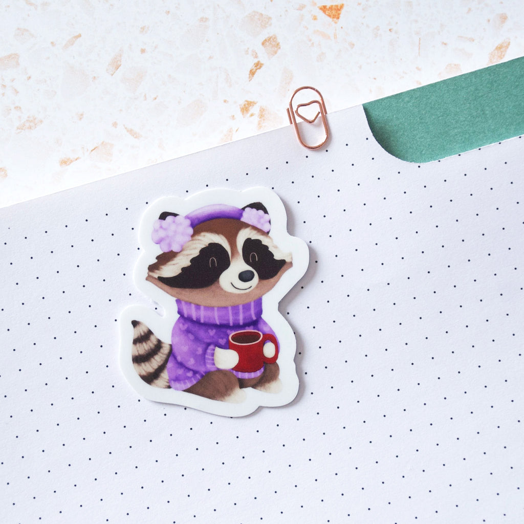 Cozy raccoon in a purple sweater, muffs and drinking cocoa vinyl sticker