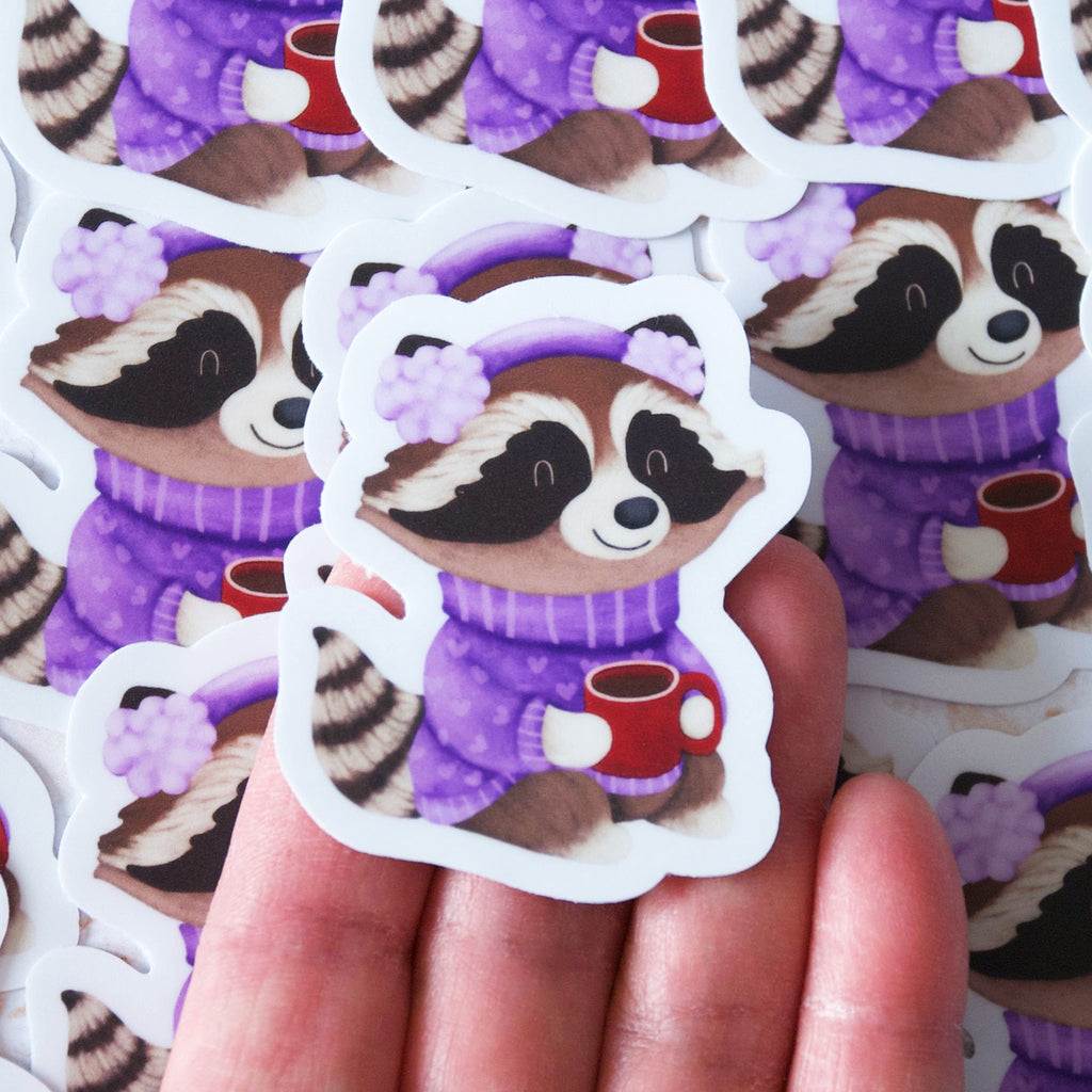 Cozy raccoon in a purple sweater, muffs and drinking cocoa vinyl sticker