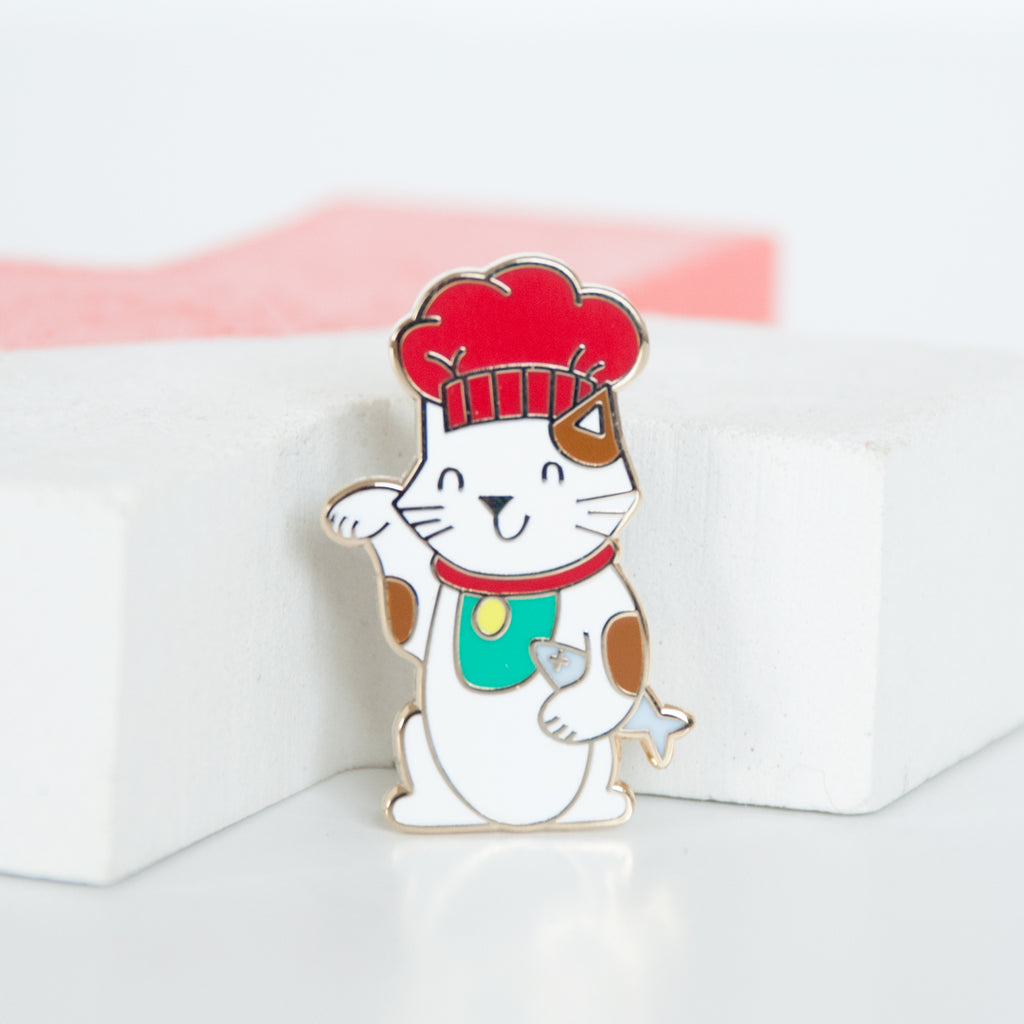 White and brown lucky cat or maneki neko cat wearing a red chef hat and holding a dead grey fish enamel pin