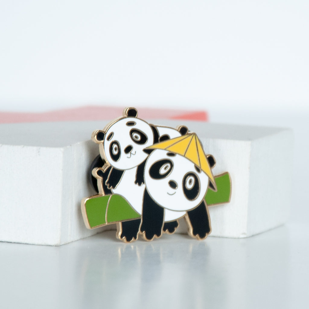 2 giant black and white pandas hanging on a bamboo branch with one panda wearing a yellow straw hat enamel pin