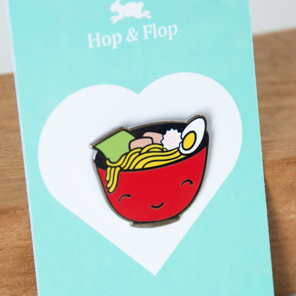 Smiling red bowl of ramen noodles enamel pin on a branded card backing