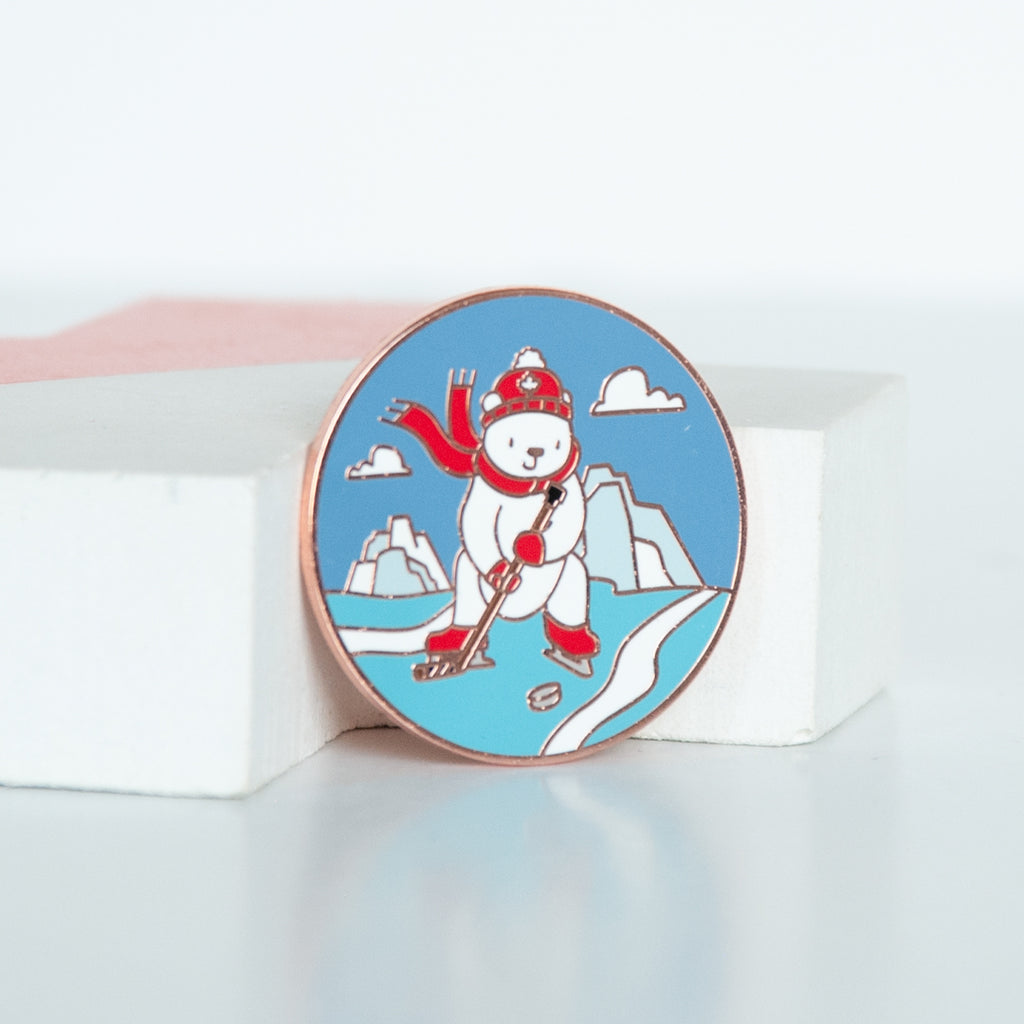 On sale white polar bear in canada coloured toque, scarf, mitts and skates playing hockey on ice with ice bergs in the background of a circle sized enamel pin