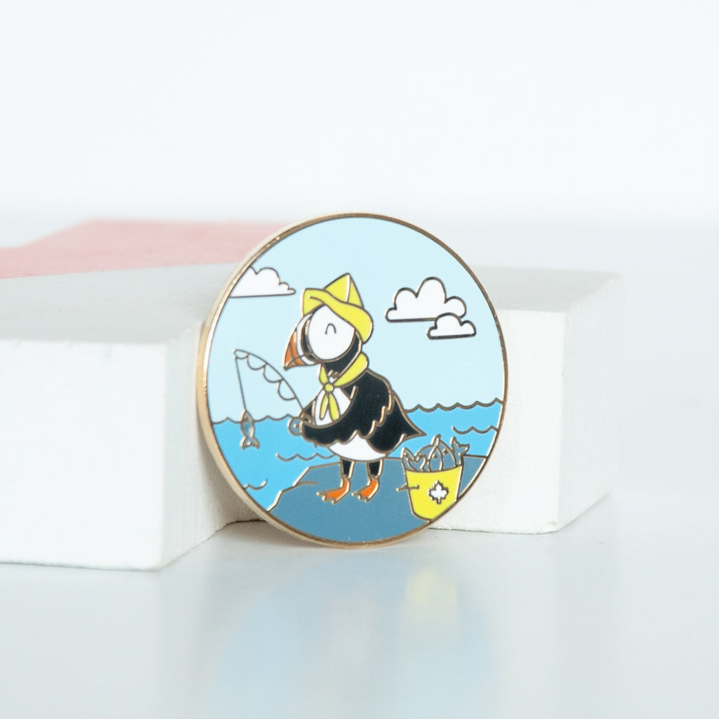 On sale east coast puffin dressed in a yellow fisherman's hat fishing in the ocean with clouds and blue sky background. A Circle hard enamel pin