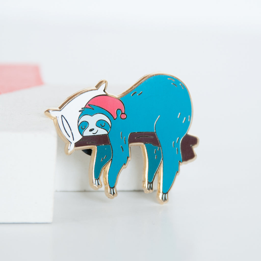 Blue sloth wearing a red cap and sleeping on a white pillow and branch enamel pin